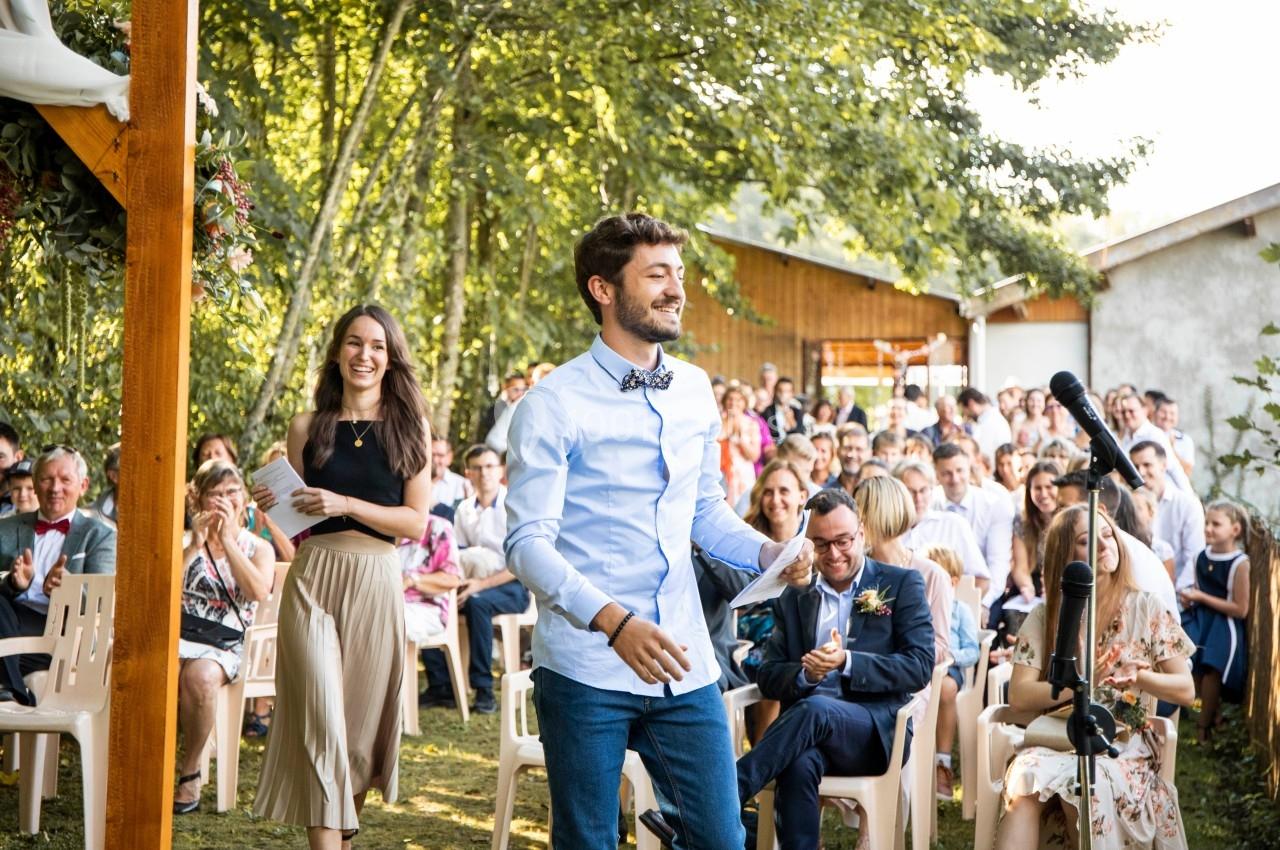 Un homme souriant marche vers un micro lors d'une cérémonie en plein air, entouré d'invités assis.