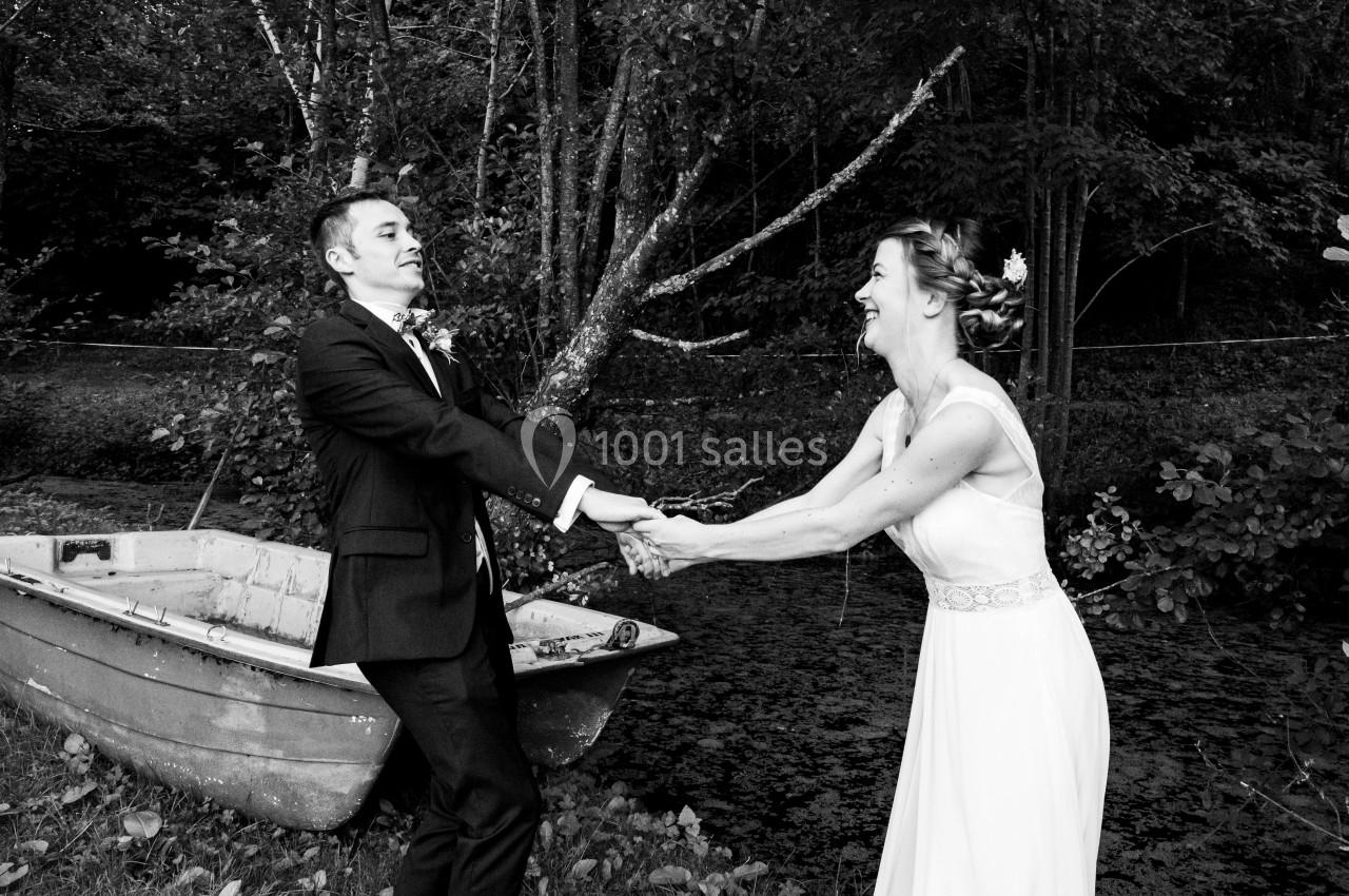 Un couple en tenue de mariage rit en se tenant les mains près d'une barque dans un cadre naturel.