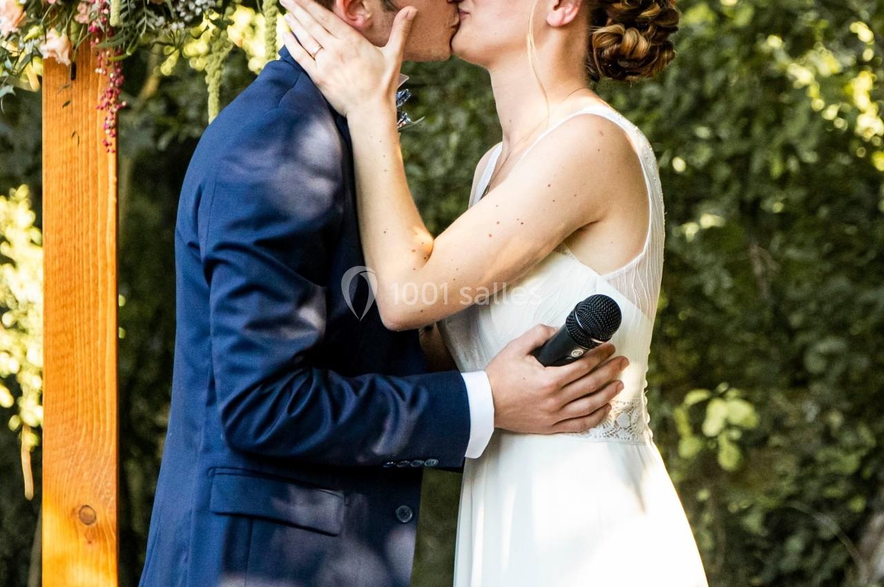 Un couple s'embrasse sous une arche décorée de fleurs lors d'une cérémonie en extérieur.