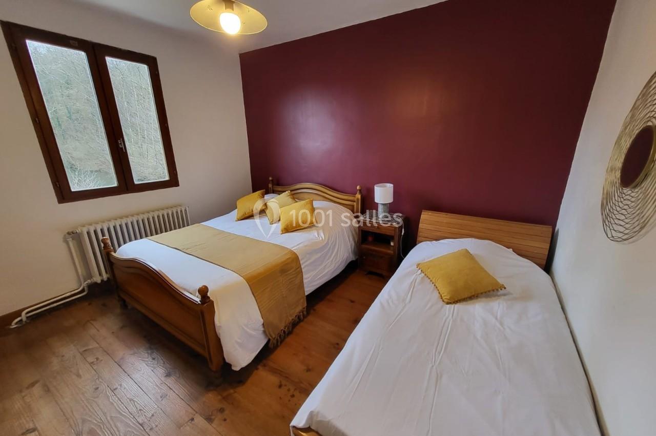 Chambre avec un lit double, un lit simple, parquet en bois, mur bordeaux et fenêtre donnant sur un extérieur verdoyant.