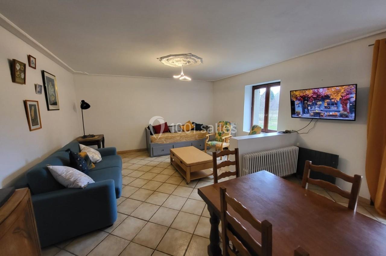 Salon lumineux avec canapé, table en bois, télévision murale, fauteuil et fenêtre donnant sur l'extérieur.