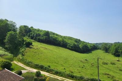 Grande maison en pierre entourée de verdure, avec une cour ensoleillée et des arbres en arrière-plan.