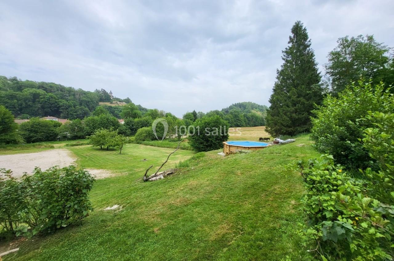 Paysage verdoyant avec pelouse, arbres, piscine hors-sol et collines boisées sous un ciel nuageux.
