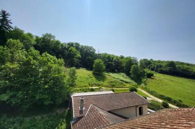 Grande maison en pierre entourée de verdure, avec une cour ensoleillée et des arbres en arrière-plan.