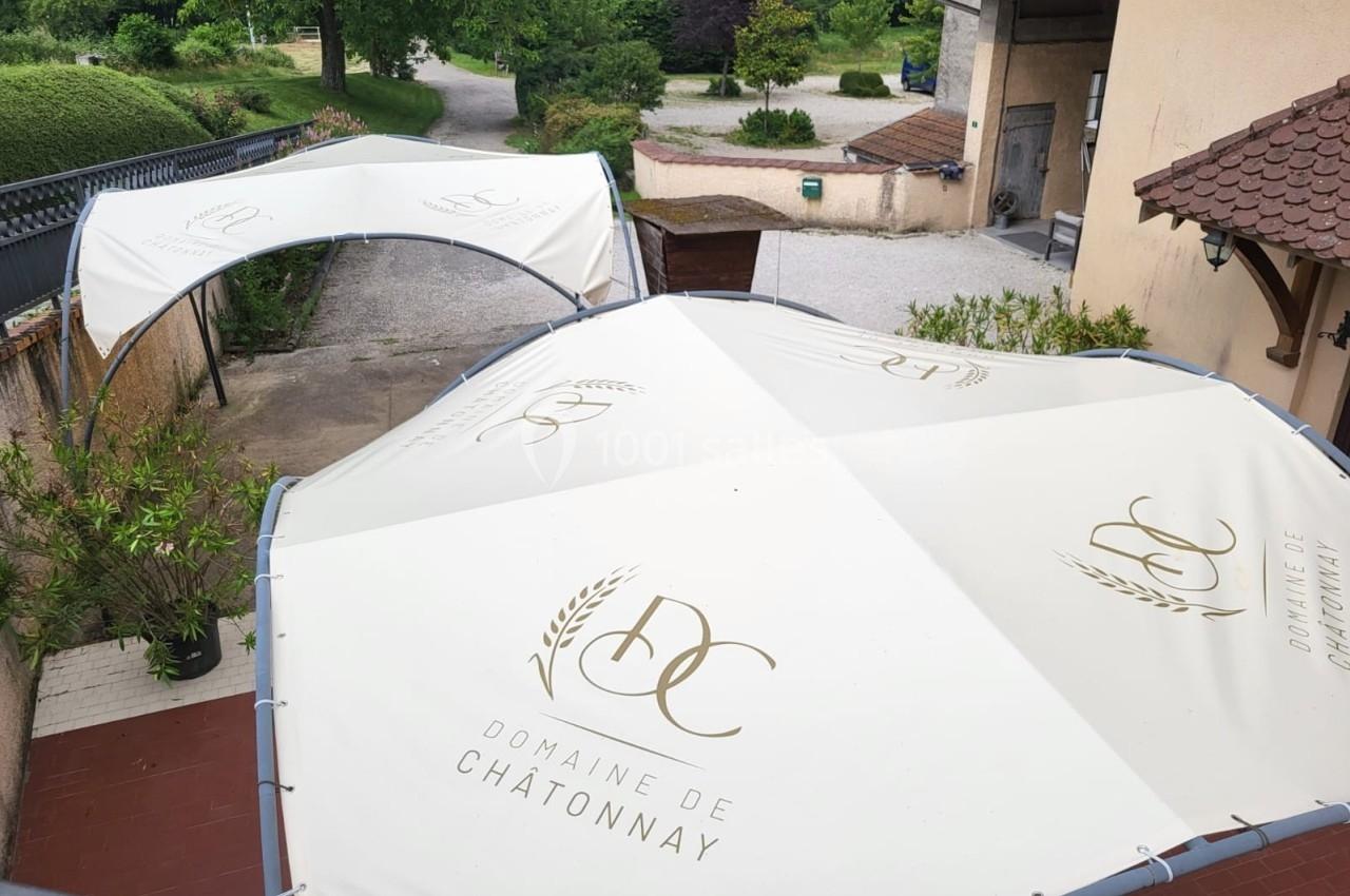 Deux grandes toiles blanches avec le logo ’Domaine de Chatonnay’ couvrent une terrasse extérieure près d'un bâtiment.