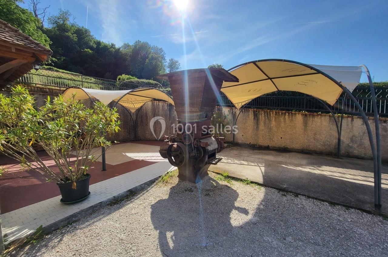 Ancien pressoir en métal exposé en extérieur sous des abris, avec un ciel ensoleillé et des arbres en arrière-plan.