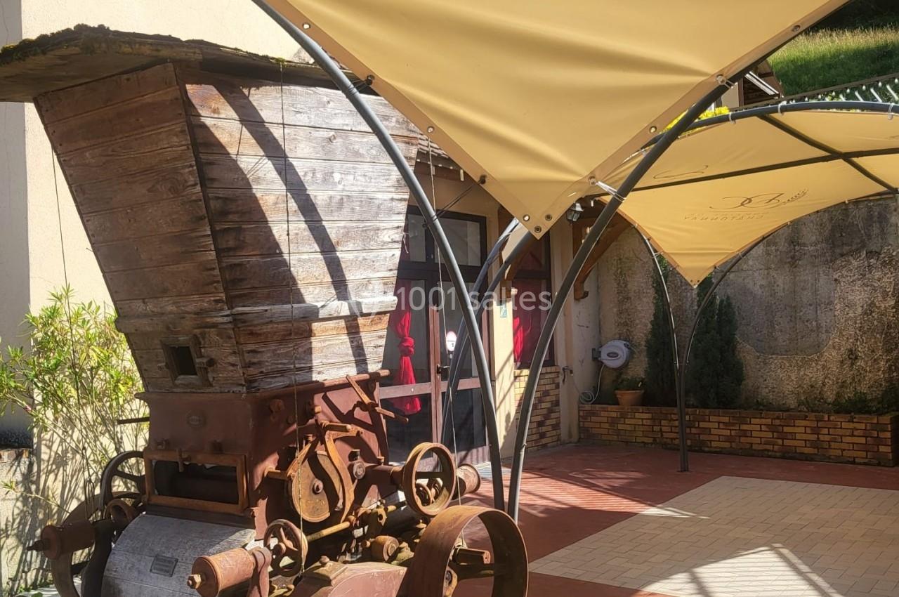 Ancien pressoir en bois et métal sous une toile tendue, situé à l'extérieur d'un bâtiment ensoleillé.