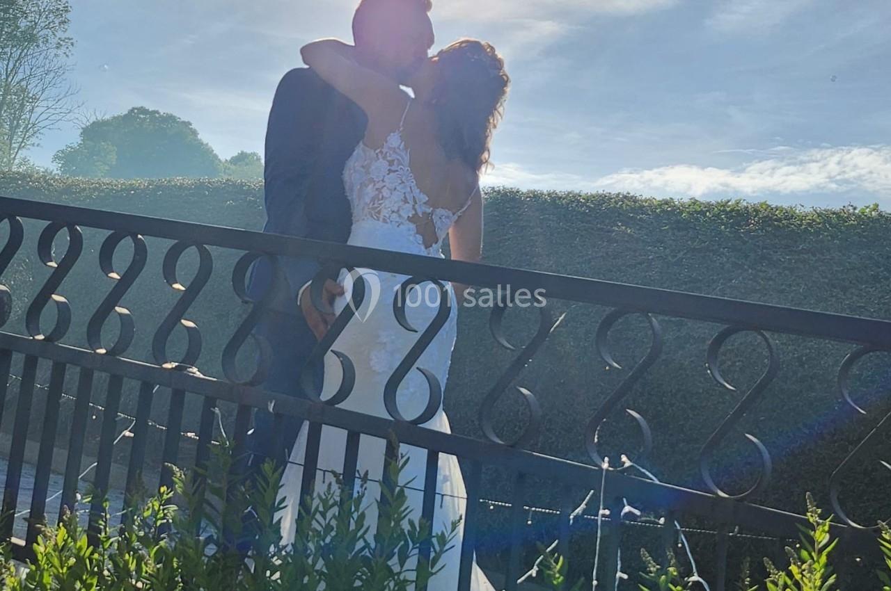 Un couple en tenue de mariage s'embrasse près d'une barrière sous un soleil éclatant.