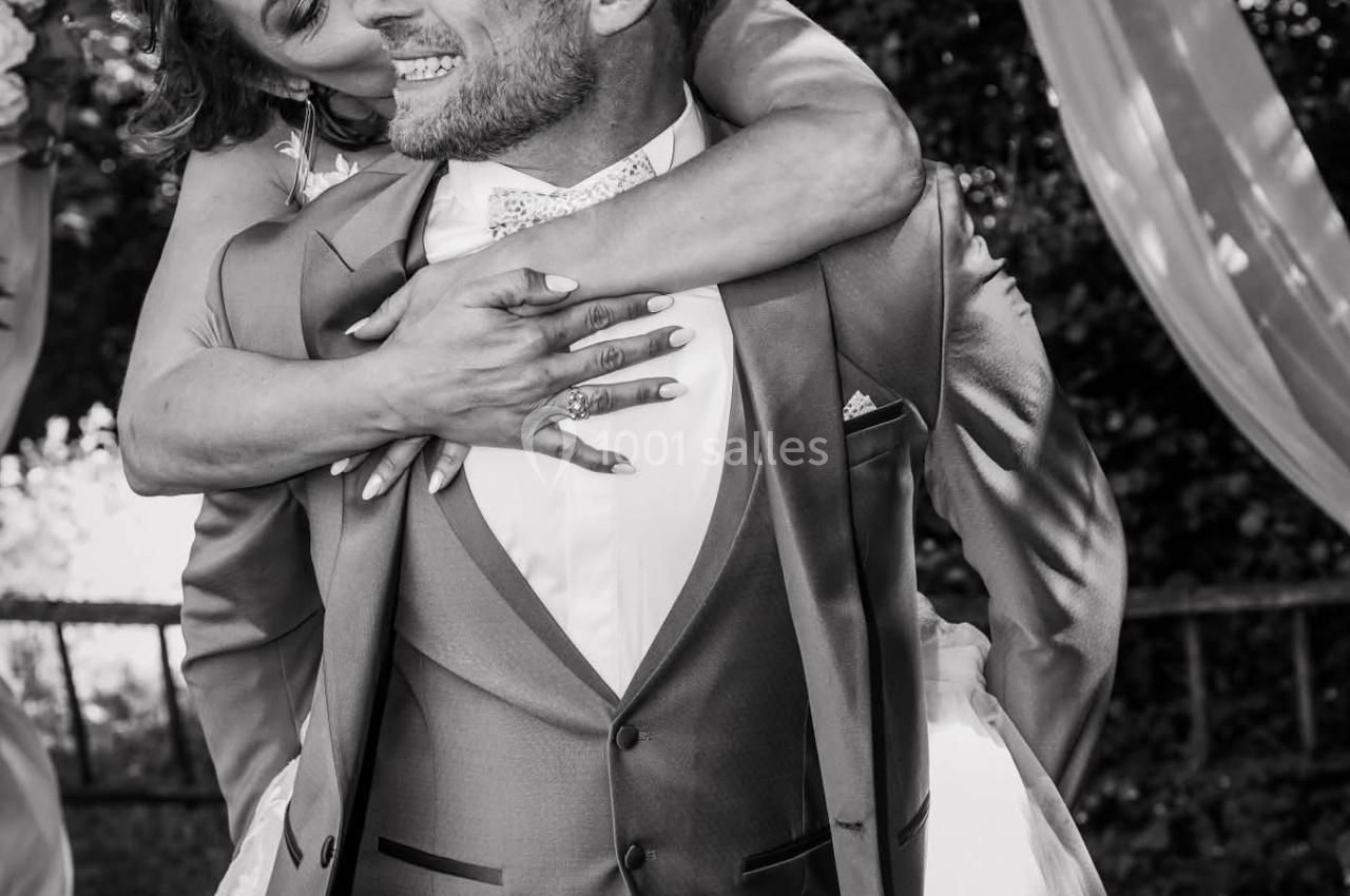 Un couple en tenue de mariage, la mariée souriante enlace le marié par derrière dans un cadre extérieur festif.