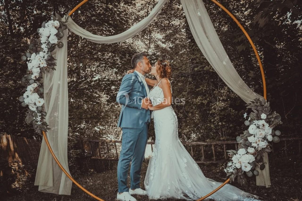 Un couple en tenue de mariage s'embrasse sous une arche décorée de fleurs dans un cadre naturel boisé.