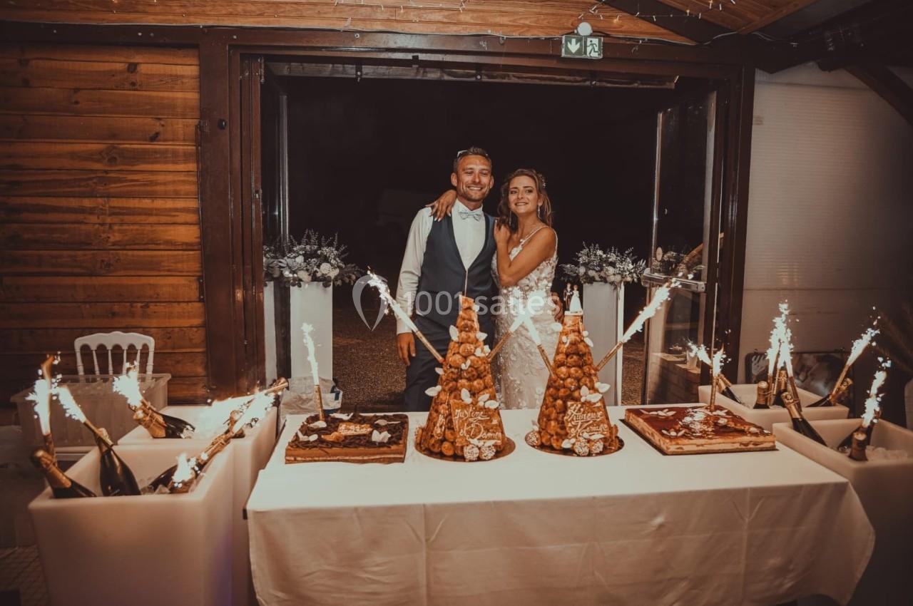 Un couple souriant pose derrière une table de desserts décorée de pièces montées et de fontaines lumineuses.