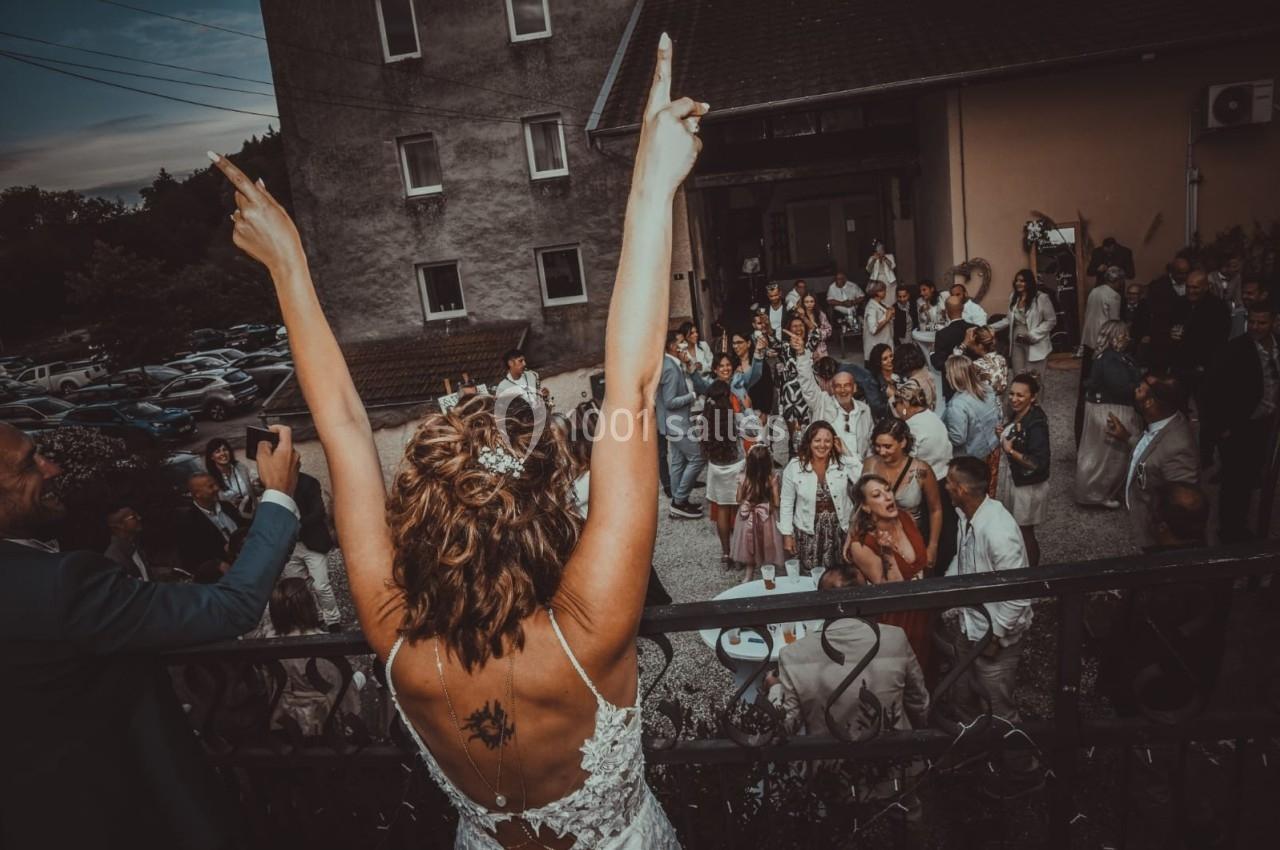 Une femme en robe de mariée lève les bras face à un groupe de personnes réunies dans une cour extérieure.