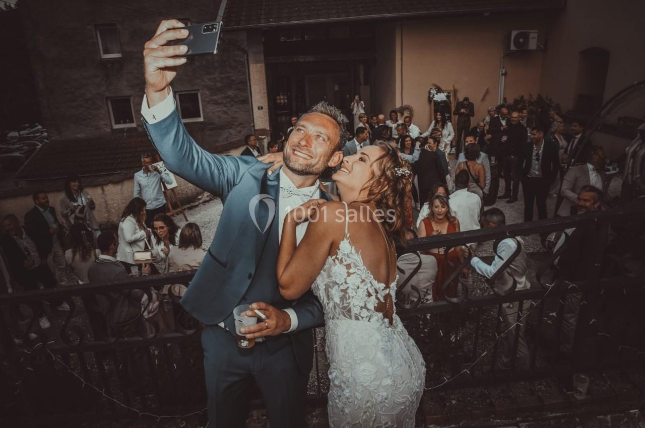 Un couple en tenue de mariage prend un selfie devant un groupe de personnes lors d'une réception en extérieur.