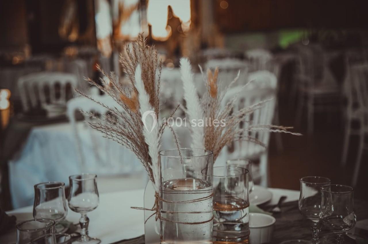 Centre de table avec des herbes séchées dans un vase en verre, entouré de verres et de vaisselle sur une table dressée.
