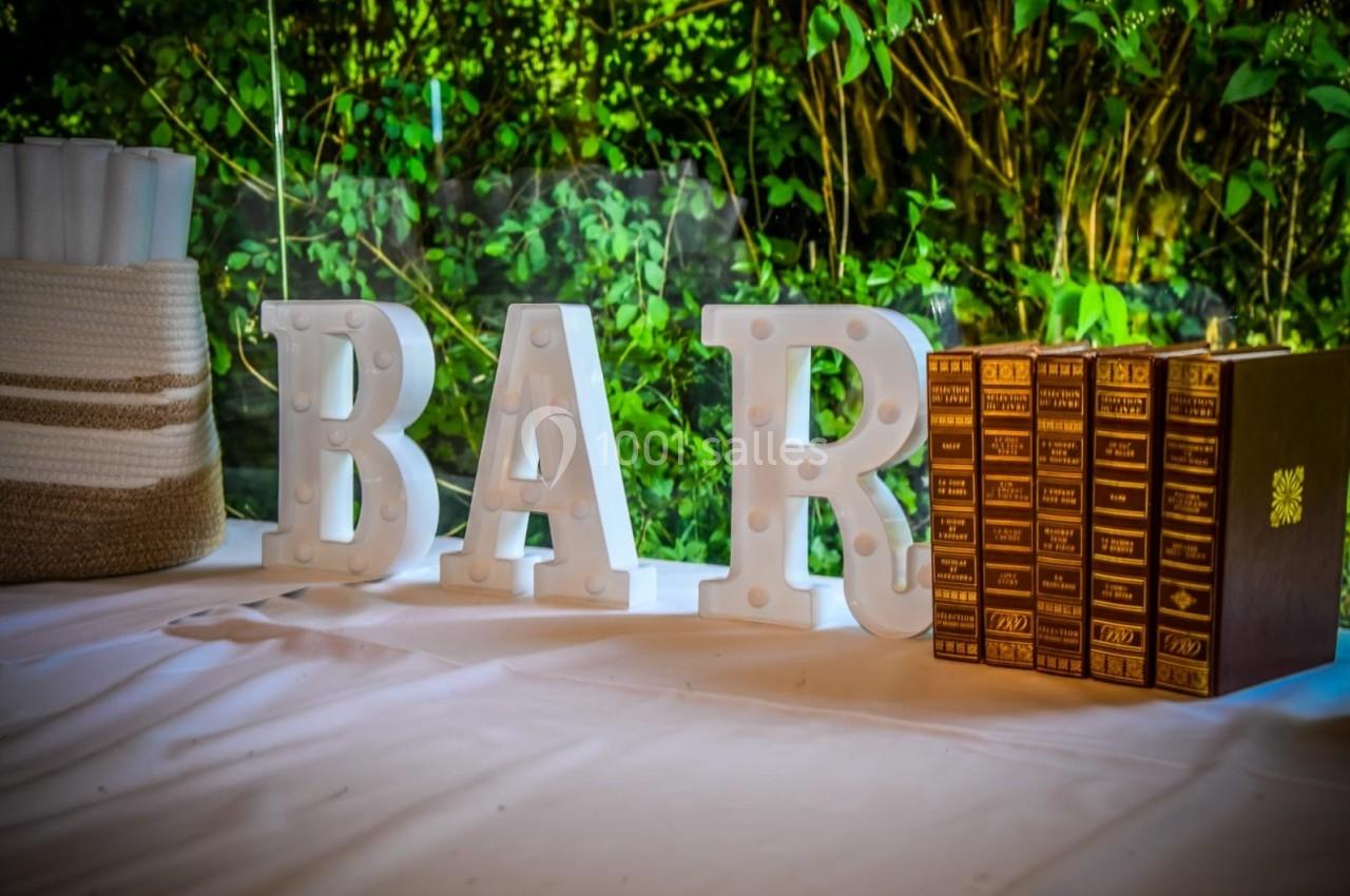 Lettres lumineuses formant ’BAR’ posées sur une nappe blanche, avec des livres dorés et un fond de végétation.