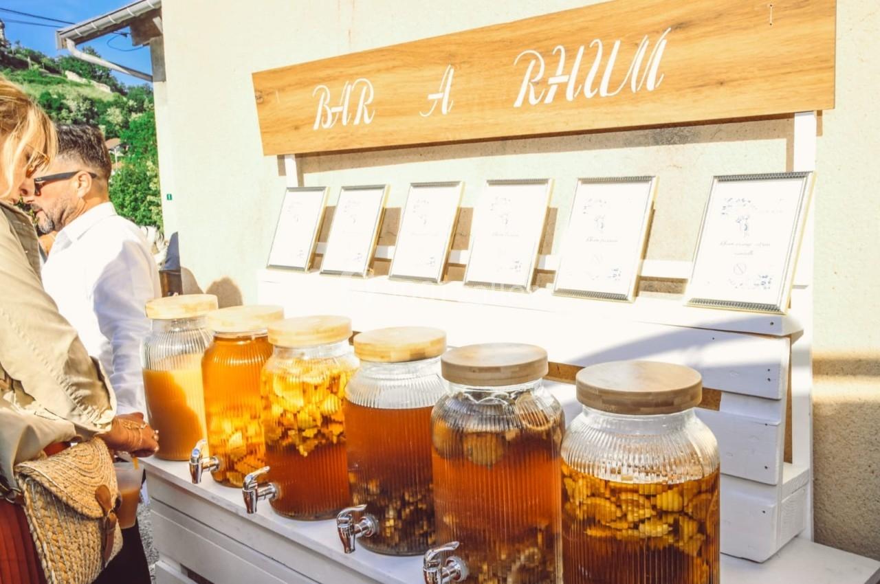 Stand avec plusieurs bocaux de rhum arrangé alignés, chacun équipé d'un robinet, sous une enseigne ’Bar à Rhum’.