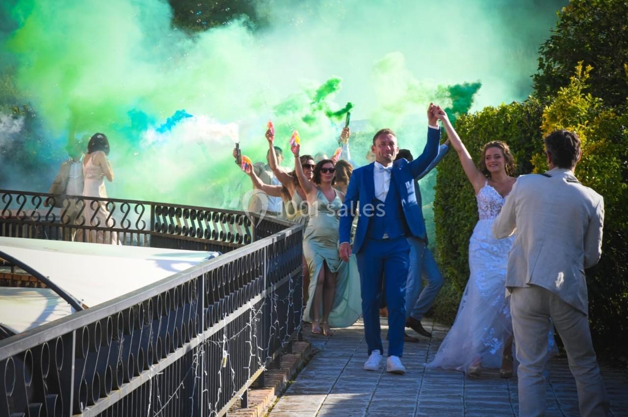 Un couple de mariés avance main dans la main, entouré d'invités brandissant des fumigènes colorés.