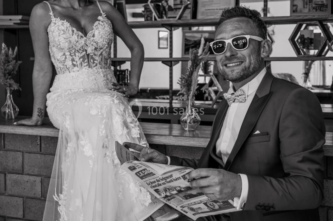 Un couple en tenue de mariage, portant des lunettes de soleil, pose de manière décontractée devant un bar.