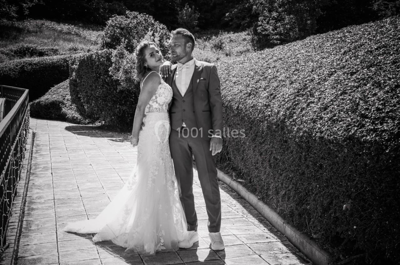 Un couple en tenue de mariage pose sur une allée pavée bordée de haies, dans un cadre extérieur ensoleillé.
