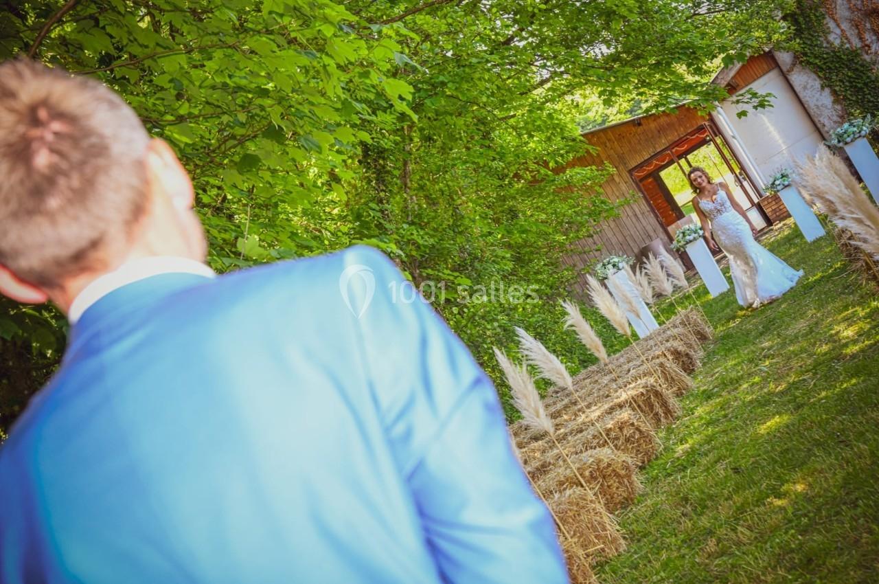 Un homme en costume bleu regarde une femme en robe de mariée marchant dans une allée bordée de bottes de foin et de plantes.
