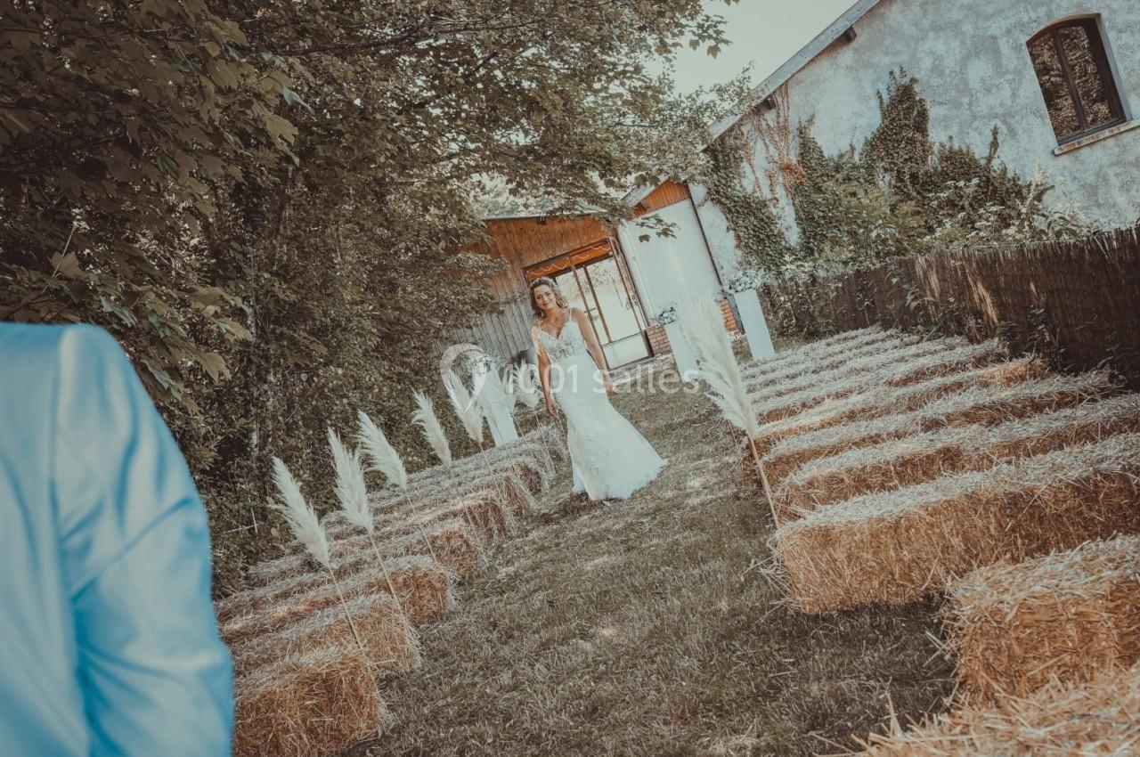 Une mariée avance dans une allée extérieure bordée de bottes de paille, entourée de verdure et de bâtiments rustiques.