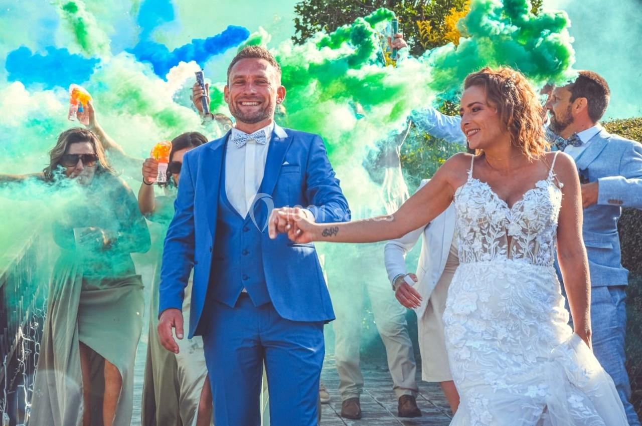 Un couple de mariés souriants marche devant des invités lançant des poudres colorées en extérieur.
