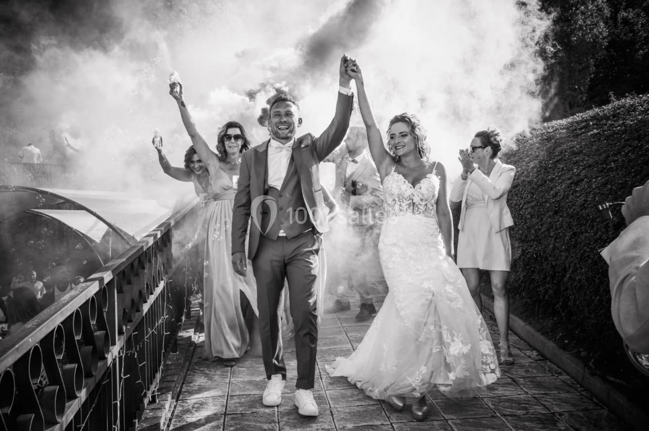 Un couple de mariés marche main dans la main, entouré d'invités et de fumée festive en extérieur.