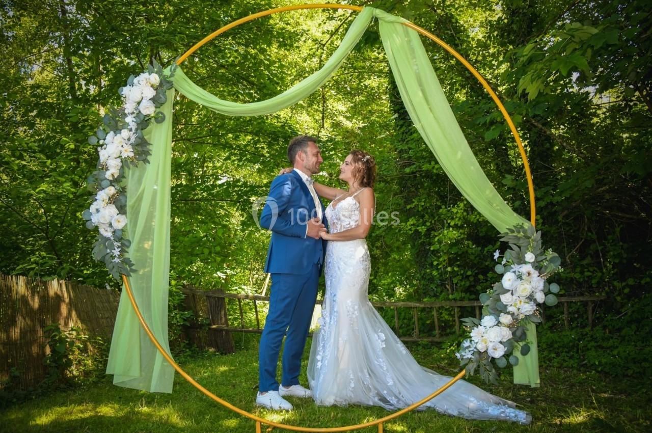 Un couple en tenue de mariage pose sous une arche circulaire décorée de fleurs blanches et de voiles verts, dans un jardin.