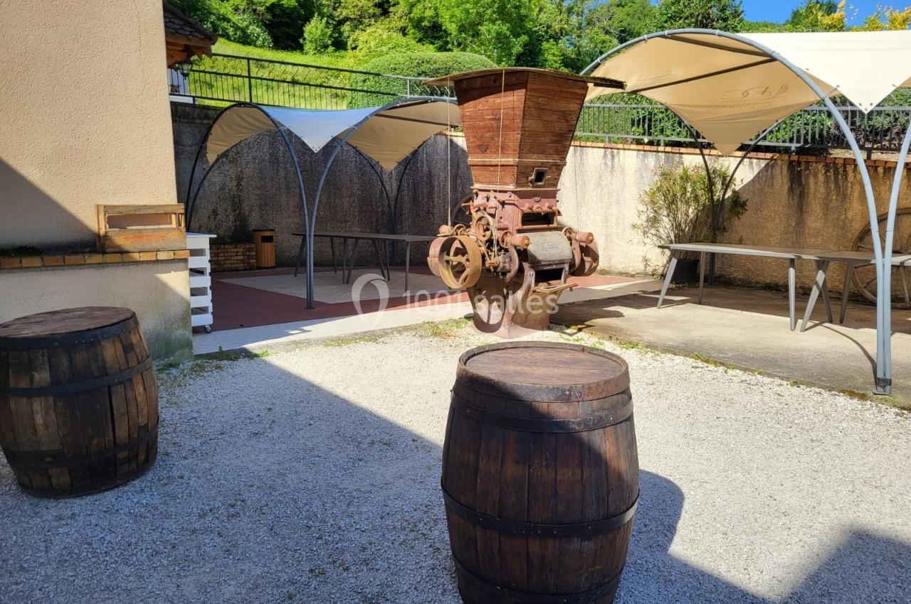 Deux tonneaux en bois devant une ancienne machine agricole sous des abris dans une cour ensoleillée.