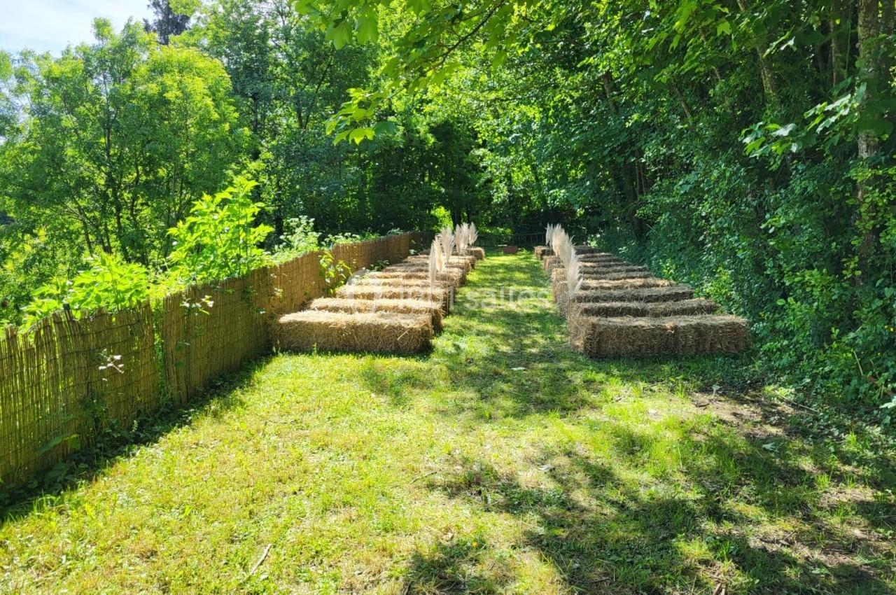 Allée enherbée bordée de bottes de paille disposées en rangées, entourée d'arbres et de verdure.