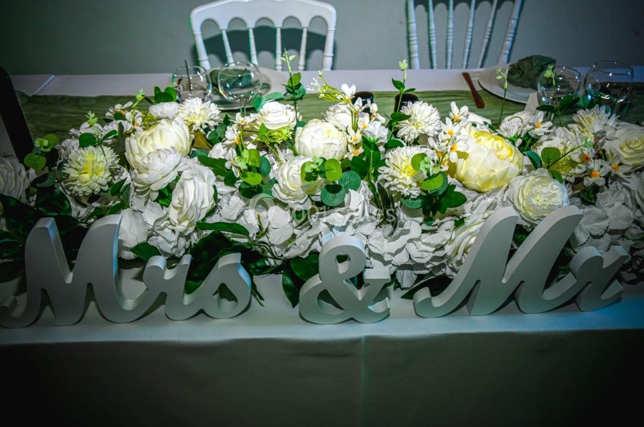 Décoration florale blanche avec feuillage vert sur une table, accompagnée des mots ’Mrs & Mr’ en lettres décoratives.