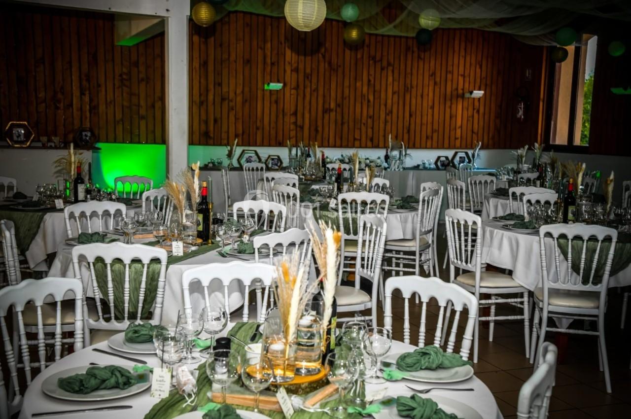 Salle de réception décorée avec des tables rondes dressées, nappes blanches, serviettes vertes et éléments naturels.