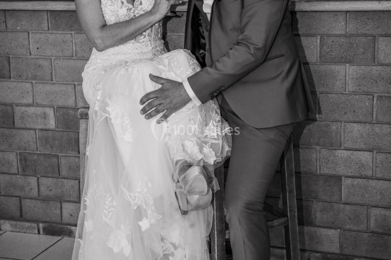 Un couple en tenue de mariage pose devant un comptoir, la mariée assise et le marié sur un tabouret.