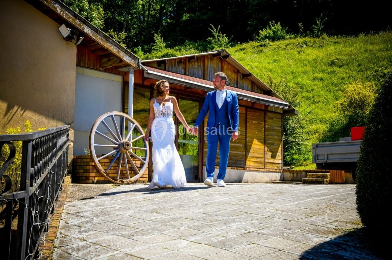 Un couple en tenue de mariage marche main dans la main devant un bâtiment en bois et une roue décorative.