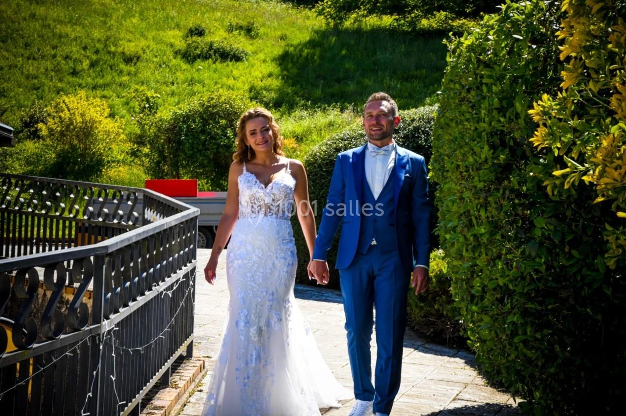 Un couple en tenue de mariage marche main dans la main sur un chemin bordé de verdure par une journée ensoleillée.