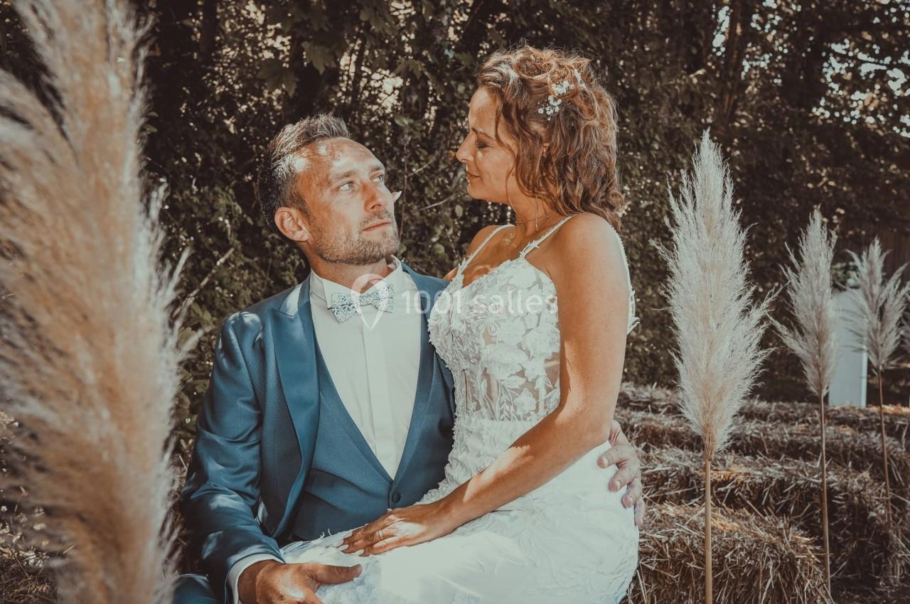 Un couple en tenue de mariage, assis sur des bottes de paille, entouré de végétation et de plumeaux décoratifs.