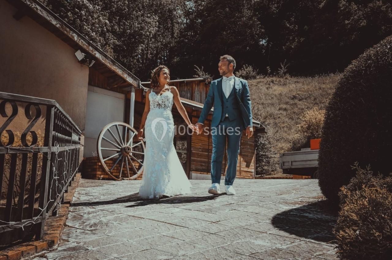 Un couple en tenue de mariage marche main dans la main sur une allée pavée, entouré de verdure et de bâtiments rustiques.
