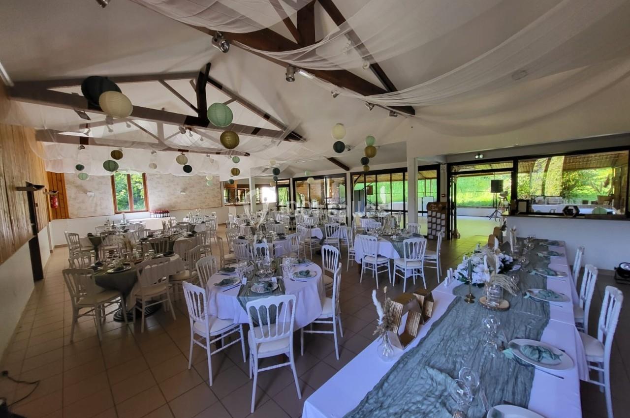 Salle de réception décorée pour un événement, avec tables dressées, chaises blanches et lanternes suspendues.