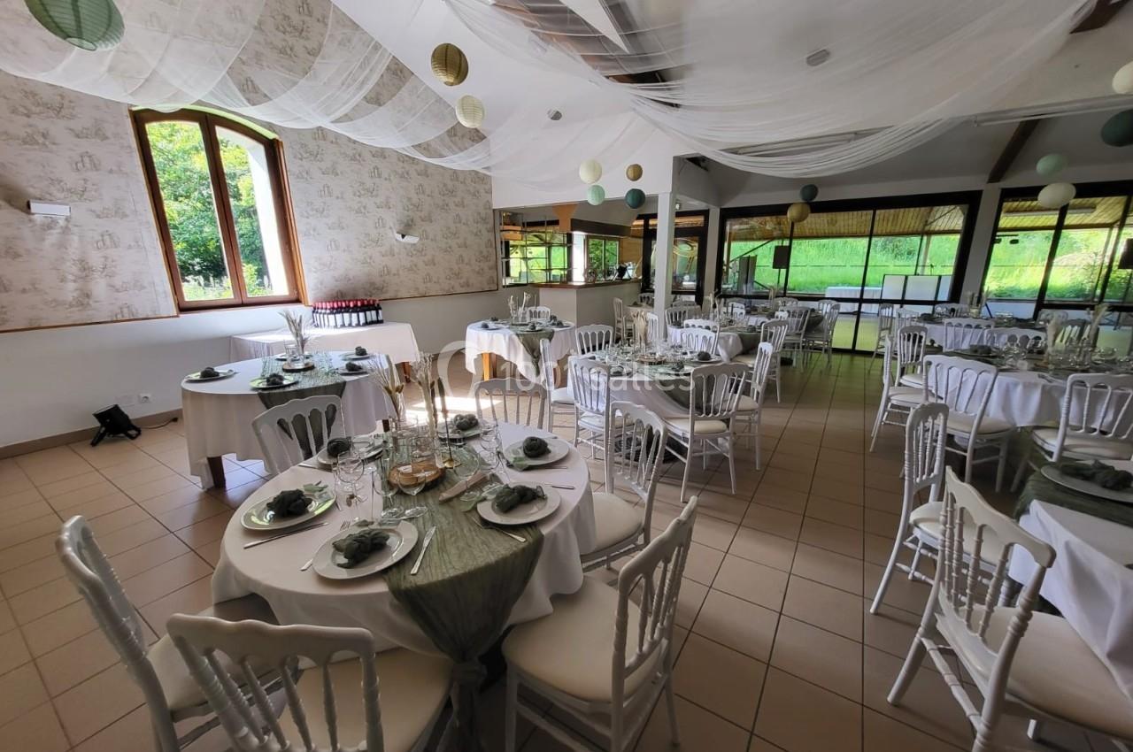 Salle de réception décorée avec des tables rondes dressées, nappes blanches et éléments verts, sous un plafond voilé.