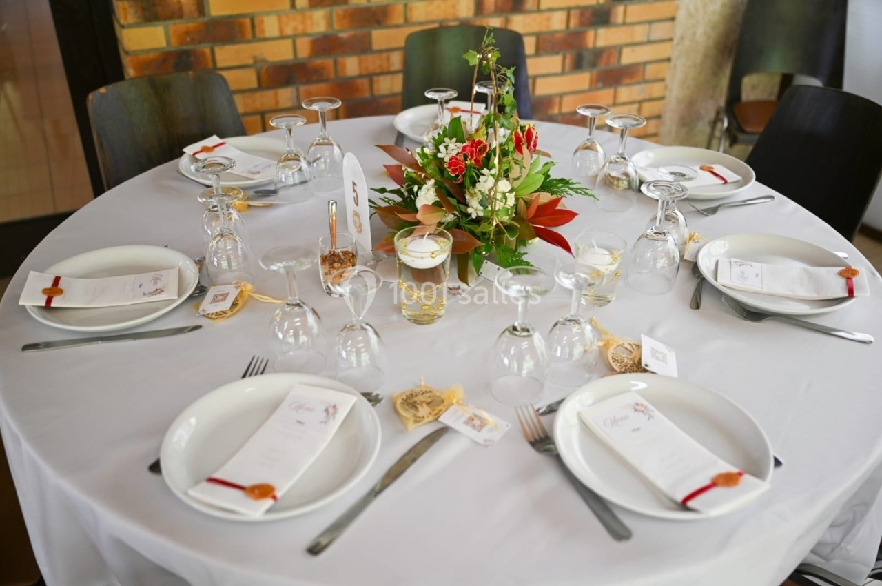 Table ronde dressée avec une nappe blanche, des assiettes, des verres, des couverts et un centre de table floral coloré.