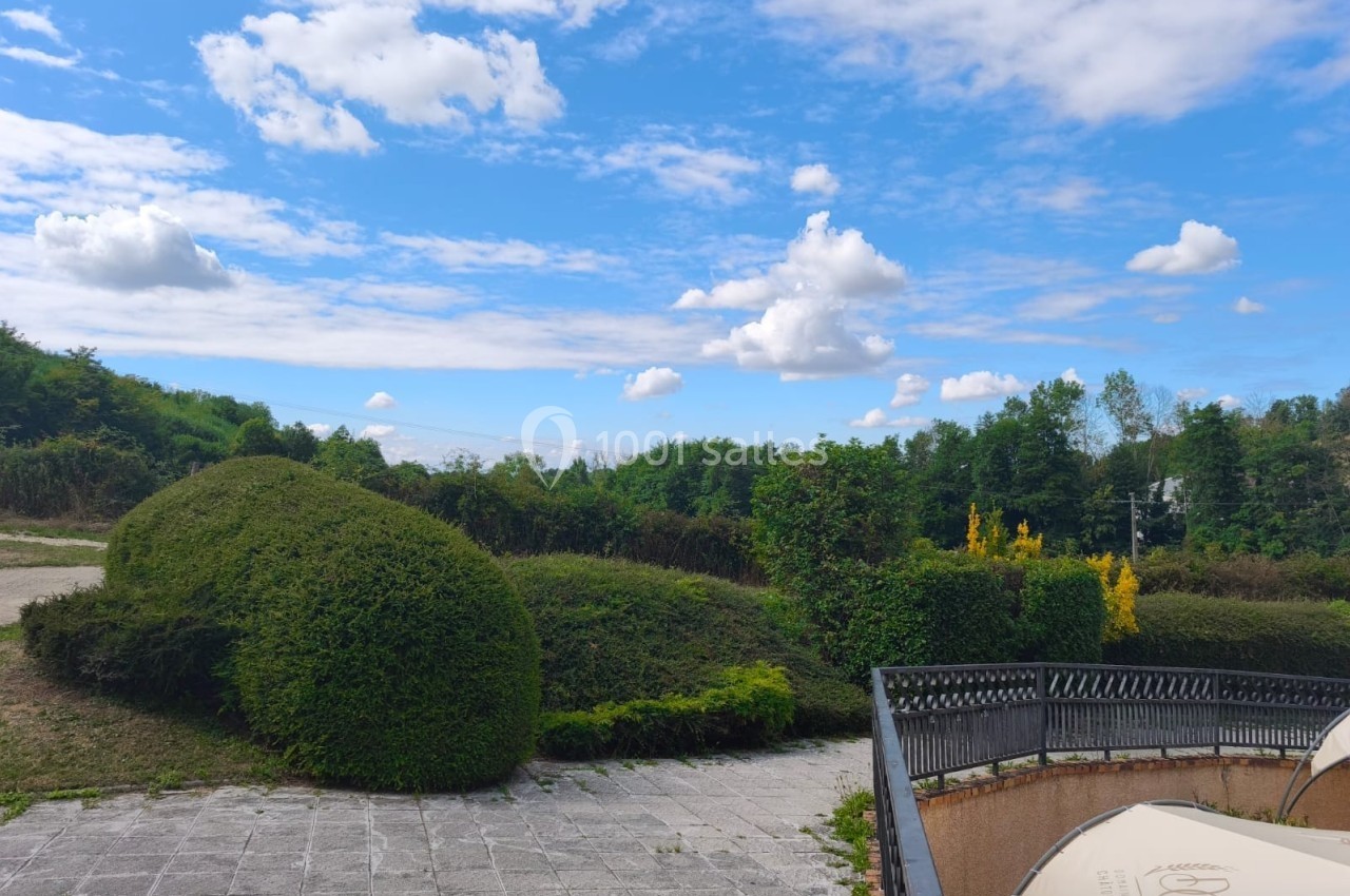 Paysage avec ciel bleu parsemé de nuages, buissons taillés, arbres verdoyants et une terrasse en premier plan.