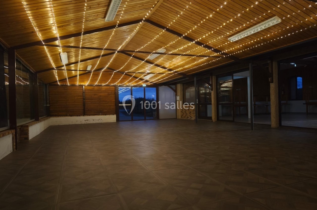 Salle vide avec plafond en bois, éclairée par des guirlandes lumineuses, grandes baies vitrées donnant sur l'extérieur.