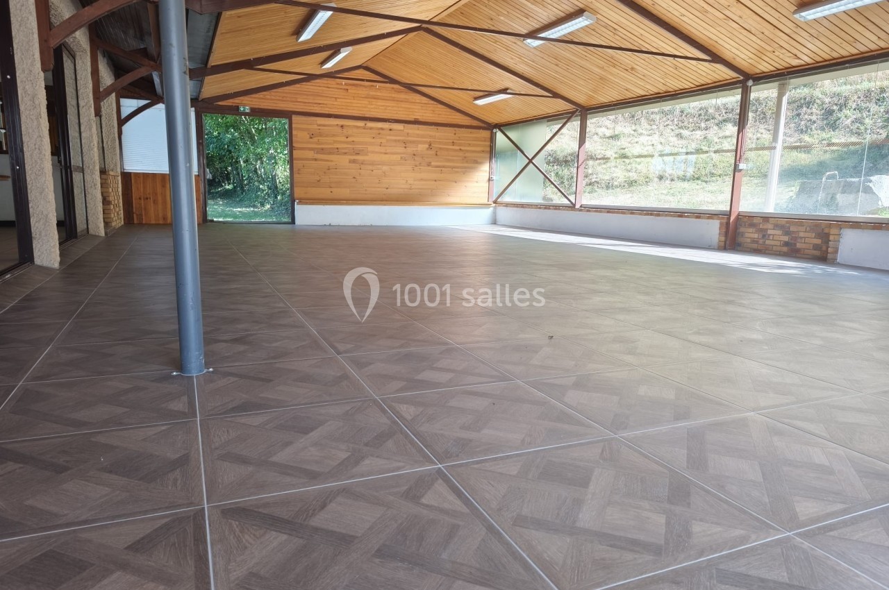Salle vide avec un sol carrelé, plafond en bois, grandes fenêtres et vue sur un espace extérieur verdoyant.