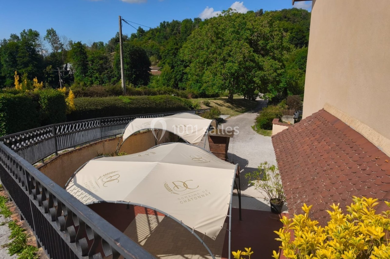Terrasse extérieure avec parasols blancs, bordée de verdure et d'une maison beige, sous un ciel dégagé.