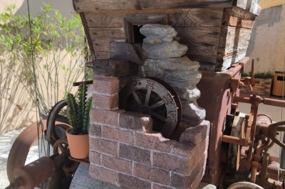 Grande maison en pierre entourée de verdure, avec une cour ensoleillée et des arbres en arrière-plan.