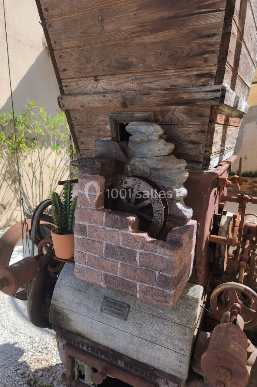 Machine agricole ancienne en bois et métal avec une roue en pierre, exposée en extérieur près d'un mur et d'une plante.