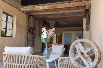 Grande maison en pierre entourée de verdure, avec une cour ensoleillée et des arbres en arrière-plan.