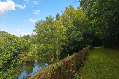 Grande maison en pierre entourée de verdure, avec une cour ensoleillée et des arbres en arrière-plan.