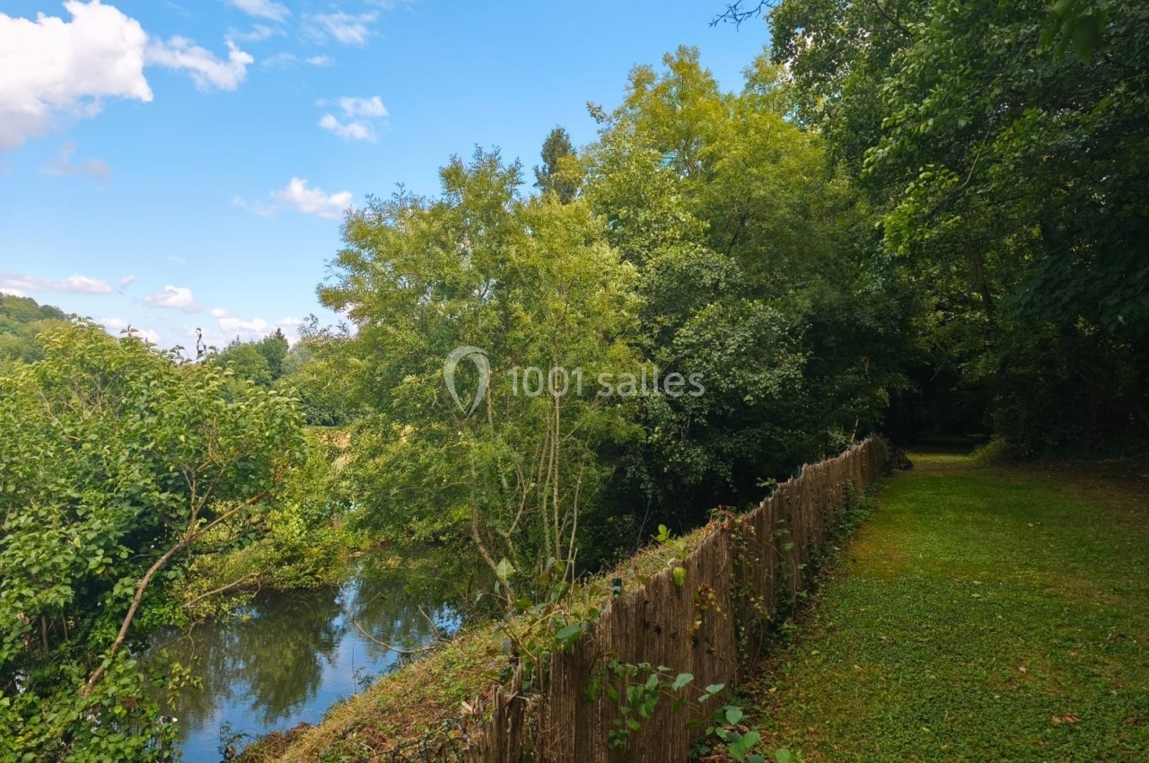 Chemin herbeux bordé d'une clôture en bois, longeant un petit cours d'eau entouré d'arbres verdoyants.