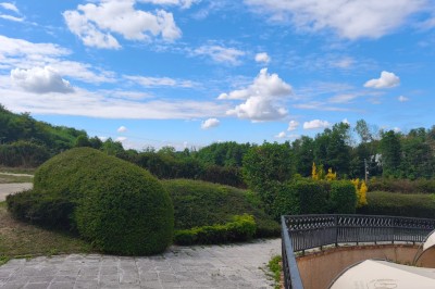 Grande maison en pierre entourée de verdure, avec une cour ensoleillée et des arbres en arrière-plan.